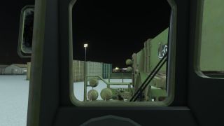 A nighttime view from inside a military vehicle cab shows green cargo containers, a floodlight, and nearby buildings under a dark sky.