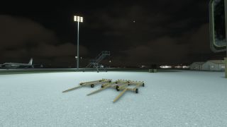 Three baggage carts sit on a lit tarmac at night, with a mobile stairway and a parked airliner in the background.