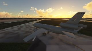 A military tri-jet aircraft sits on a wet taxiway at sunrise, with scattered clouds and runways visible in the background.