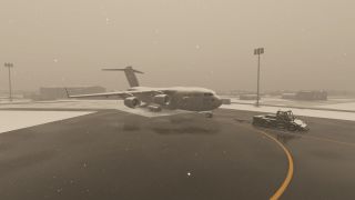 A large military cargo aircraft is being towed by a pushback tug on a snow-covered airport ramp during active snowfall.