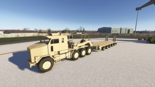 A tan military-style heavy transport truck with a multi-axle flatbed trailer is parked on a concrete surface near hangars and fencing.