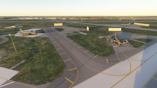 Several large military cargo aircraft are parked on concrete aprons near taxiways, with grassy areas and blast walls visible nearby.