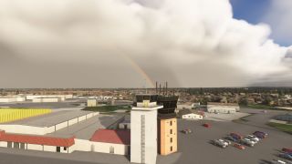 A control tower stands beside several hangars and a parking lot, with a faint rainbow visible under overcast skies in the background.