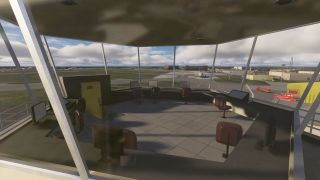 An airport control tower interior features empty chairs and computer monitors overlooking taxiways, a parked aircraft, and red airport vehicles outside.