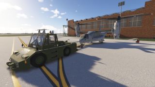 A military green tow tractor is pulling a tiltrotor aircraft on a sunny ramp beside a large brick hangar.
