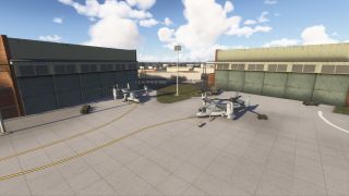 Two tiltrotor aircraft are parked on a concrete apron between large green hangars, with ground vehicles and equipment nearby under a partly cloudy sky.