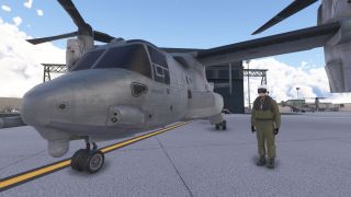 A tiltrotor military aircraft is parked on a ramp with its rotors level, while a pilot in a green flight suit stands nearby.