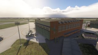 A large brick hangar with green doors stands beside a ramp where several military tiltrotor aircraft are parked under cloudy skies.
