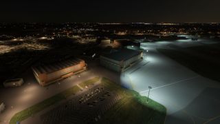 Several large hangars and a parking lot are illuminated by floodlights at an airport apron during nighttime.