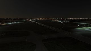 A long runway with illuminated edge lights stretches into the distance at night, surrounded by taxiways and apron areas with floodlights.