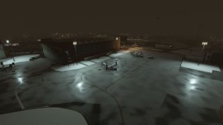 A snowy airport apron at night is illuminated by floodlights, with two tiltrotor aircraft parked near a large hangar building.