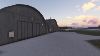 Two large, curved-roof hangars stand in the foreground while several military aircraft are parked on the apron at sunset.