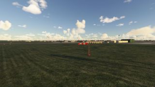 A red and white windsock stands on a grassy field near several airport buildings under a partly cloudy sky.