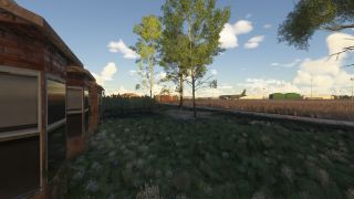 A brick building with dark windows stands beside tall grass and trees, with a military transport aircraft visible near distant hangars.