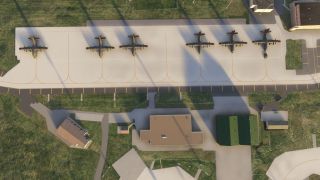 Six military transport aircraft are parked in a row on a concrete apron beside several buildings and grassy areas viewed from above.