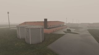 A large rectangular building with a red exterior sits beside two cylindrical tanks and a small utility trailer on a wet, overcast airfield.