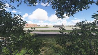 A military jet marked "U.S. Air Force" sits on a taxiway, partially viewed through leafy bushes with buildings in the background.