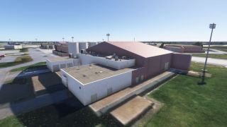 A large hangar with rooftop HVAC units and adjacent utility buildings sits beside a taxiway under clear blue skies.