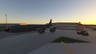 A dark gray U.S. Air Force jet with four engines is parked on a ramp near a hangar at sunrise, with a military truck nearby.