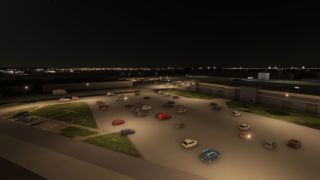 A large parking lot filled with various cars is illuminated by overhead lights beside several hangar buildings at night.