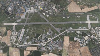Aerial view shows a large airport with a single long runway, multiple taxiways, parked aircraft, hangars, and surrounding fields and buildings.
