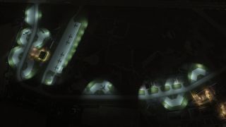 Several military aircraft are parked on illuminated aprons at night, surrounded by taxiways, hangars, and scattered buildings in a darkened airfield.