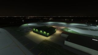 A nighttime airport scene shows green-lit hangars, a parked military aircraft on a ramp, and distant city lights on the horizon.