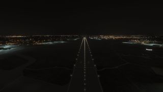 A runway is illuminated by edge and centerline lights at night, with airport buildings and city lights visible in the distance.