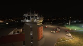 An airport control tower stands illuminated at night with several parked vehicles and city lights visible in the distant background.