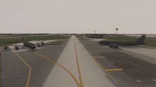 Two large gray military aircraft are parked on adjacent taxiways at an airfield, with additional aircraft and buildings visible in the background.