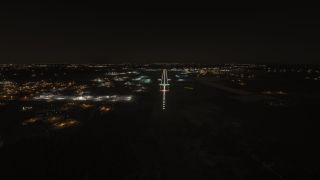 A runway with bright approach and edge lights is seen at night, surrounded by illuminated airport buildings and distant city lights.