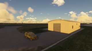 A tan utility vehicle is parked on a paved area beside a small beige hangar, with scattered clouds and airport infrastructure in the distance.