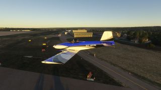A blue and white aerobatic aircraft marked EC-KCN flies low over airport taxiways, with hangars and trees behind it.