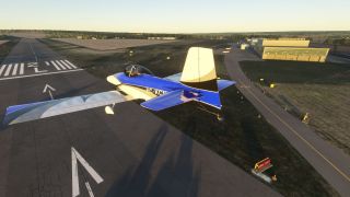 A blue and white single-engine aircraft flies low over runway threshold markings, with a taxiway and hangar to the right.