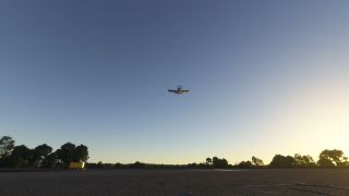 A single-engine propeller aircraft flies above a paved airfield, with trees lining the horizon and warm sunlight brightening the clear sky.
