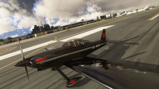 A black RV-4 with red graphics and two occupants sits on airport pavement, with terminal buildings and airliners behind it.