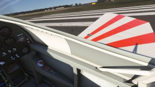The cockpit view shows analog gauges, toggle switches, and a red-and-white striped wing beside white runway markings.