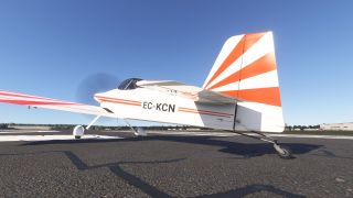 A white tailwheel aircraft with orange sunburst markings and registration EC-KCN sits on a runway with its propeller spinning.