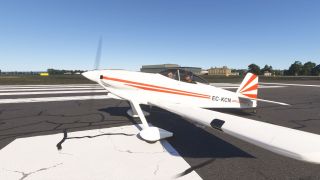 A white low-wing aircraft with orange stripes and two occupants sits on runway markings with its propeller spinning.