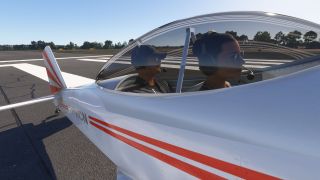 Two headset-wearing occupants sit beneath a bubble canopy in a white aircraft with red stripes beside runway markings.