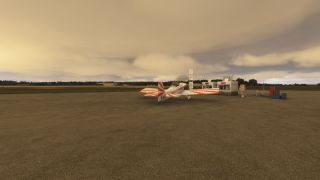A red-and-white light aircraft sits beside airport fuel pumps on a broad apron beneath low, layered clouds.
