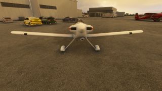A white low-wing aircraft faces forward on the apron, with parked cars beside hangars and a red biplane to the right.