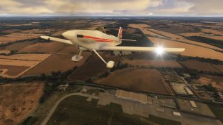 A white low-wing aircraft with red stripes and fixed landing gear flies over patchwork fields and industrial buildings beneath cloudy skies.
