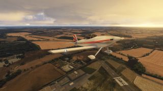 A white low-wing aircraft with red stripes and fixed landing gear flies above a rural runway and patchwork fields beneath golden clouds.