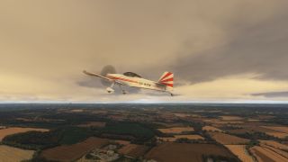 A white propeller aircraft with red stripes and fixed landing gear flies above patchwork farmland beneath low clouds.