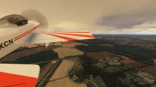 A white propeller aircraft with red wing stripes flies above a runway, patchwork fields, and wooded areas beneath an overcast sky.