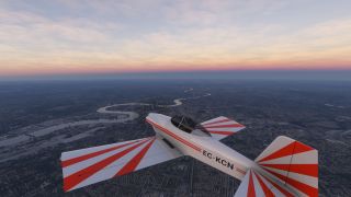A white aircraft with red sunburst markings flies above a dense city and winding river beneath a pink-tinged sky.