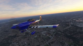 A blue and silver aircraft with striped wings and fixed landing gear flies above dense neighborhoods beneath a yellow-orange horizon.