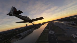 A black single-engine aircraft with a checkered tail banks above runway 09 beside a waterway under an orange evening sky.