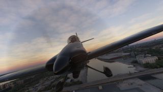 A dark single-engine airplane with fixed landing gear banks above a city river and bridge beneath a cloudy sunset sky.